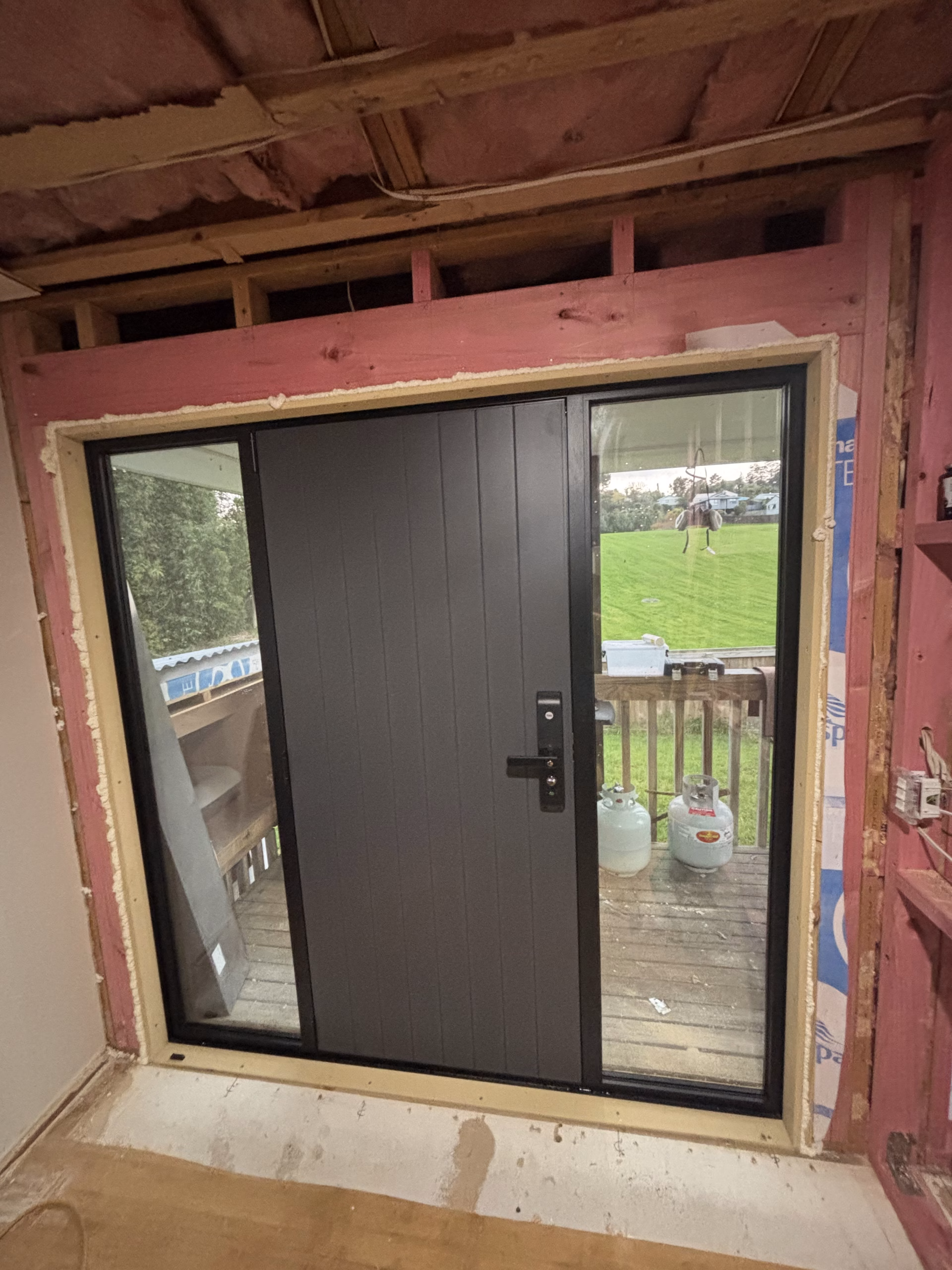 Dark grey aluminium entry door with side glass panels, freshly installed in a timber-framed opening during a home renovation