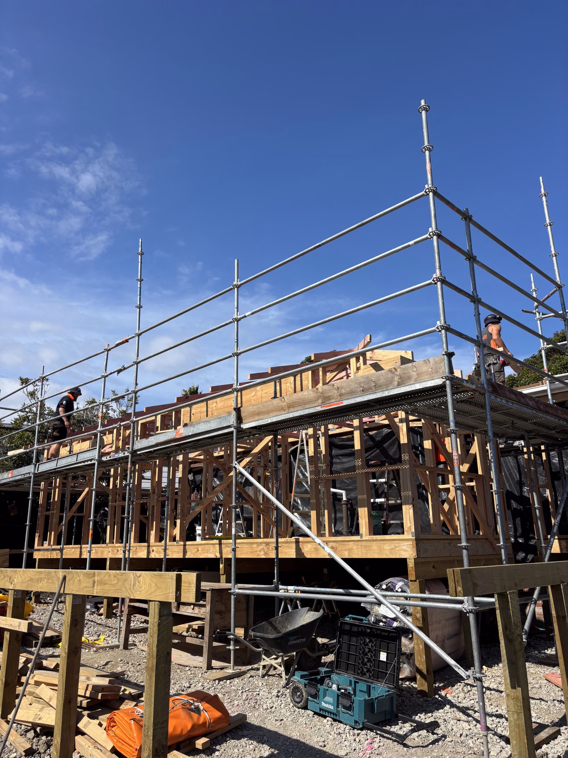 House under construction with timber framing and scaffolding, two workers on site, tools and materials on the ground below