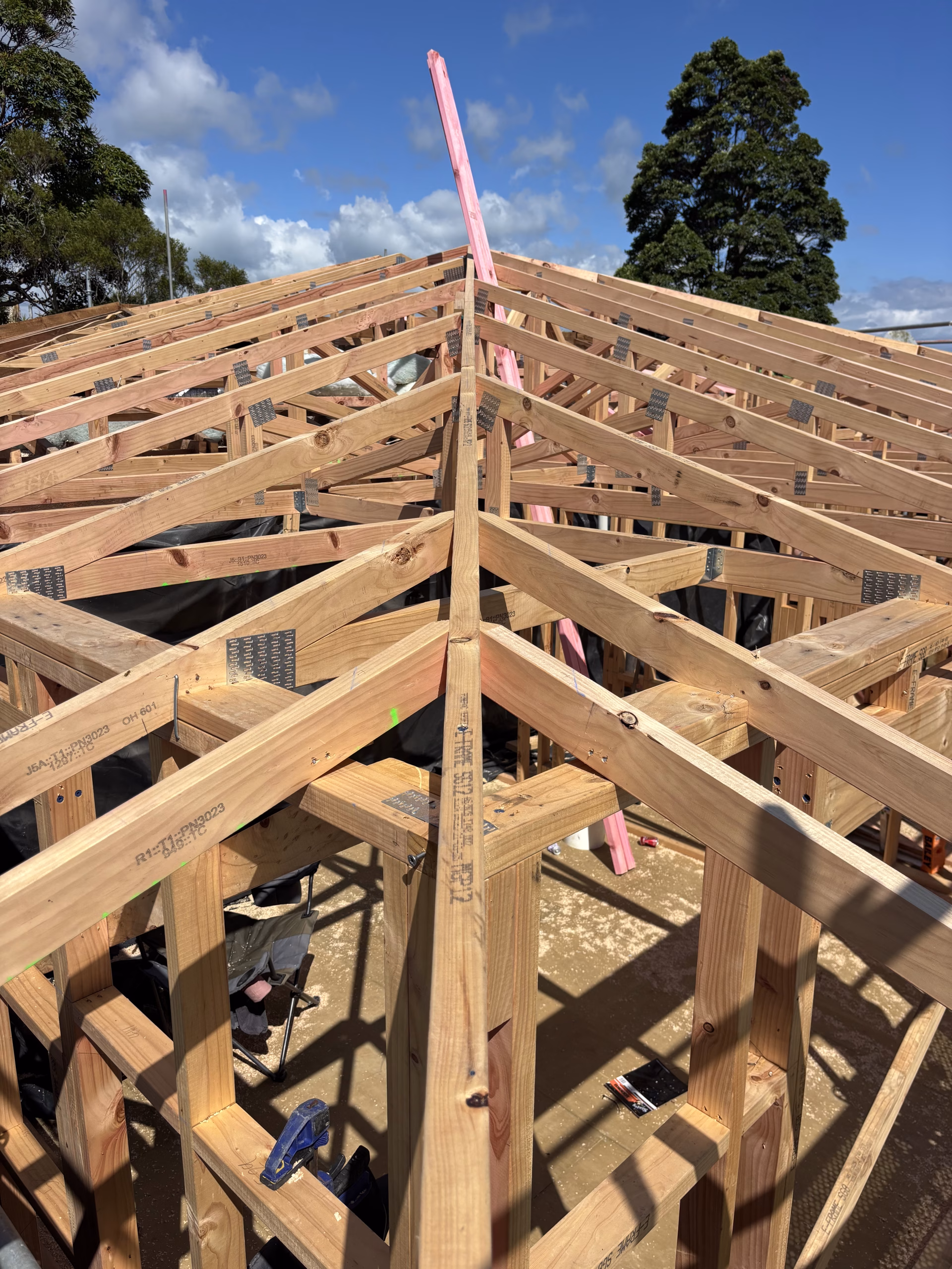 Timber roof framing under construction, showing rafters meeting at a central ridge with metal nail plates and bracing visible
