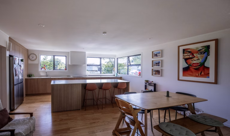Open-plan kitchen and dining area with timber cabinetry, island bench, bar stools, and large windows overlooking a garden