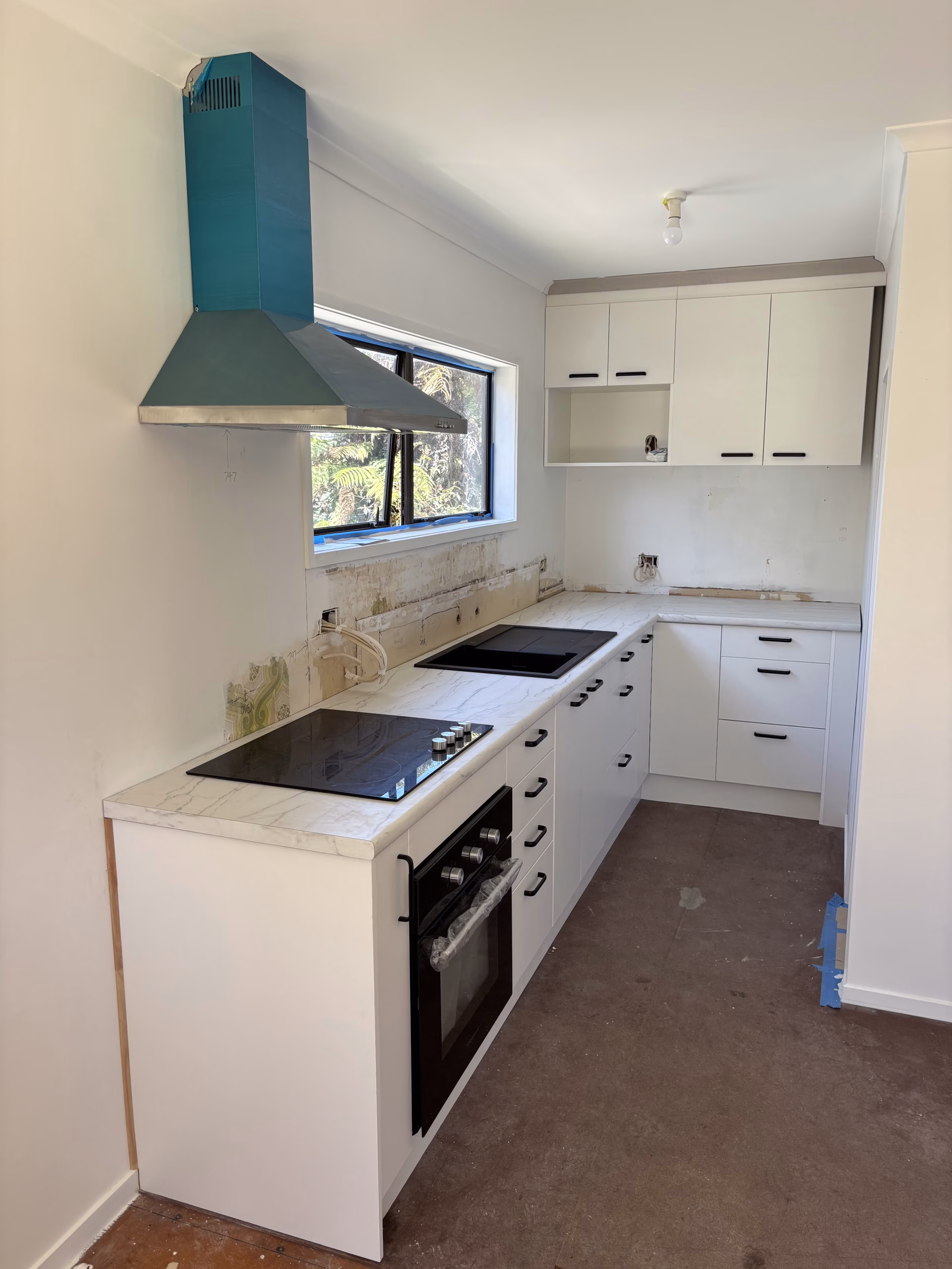 Kitchen renovation in progress with white cabinetry, marble-look benchtops, black appliances, and a teal rangehood, with flooring and splashback yet to be completed