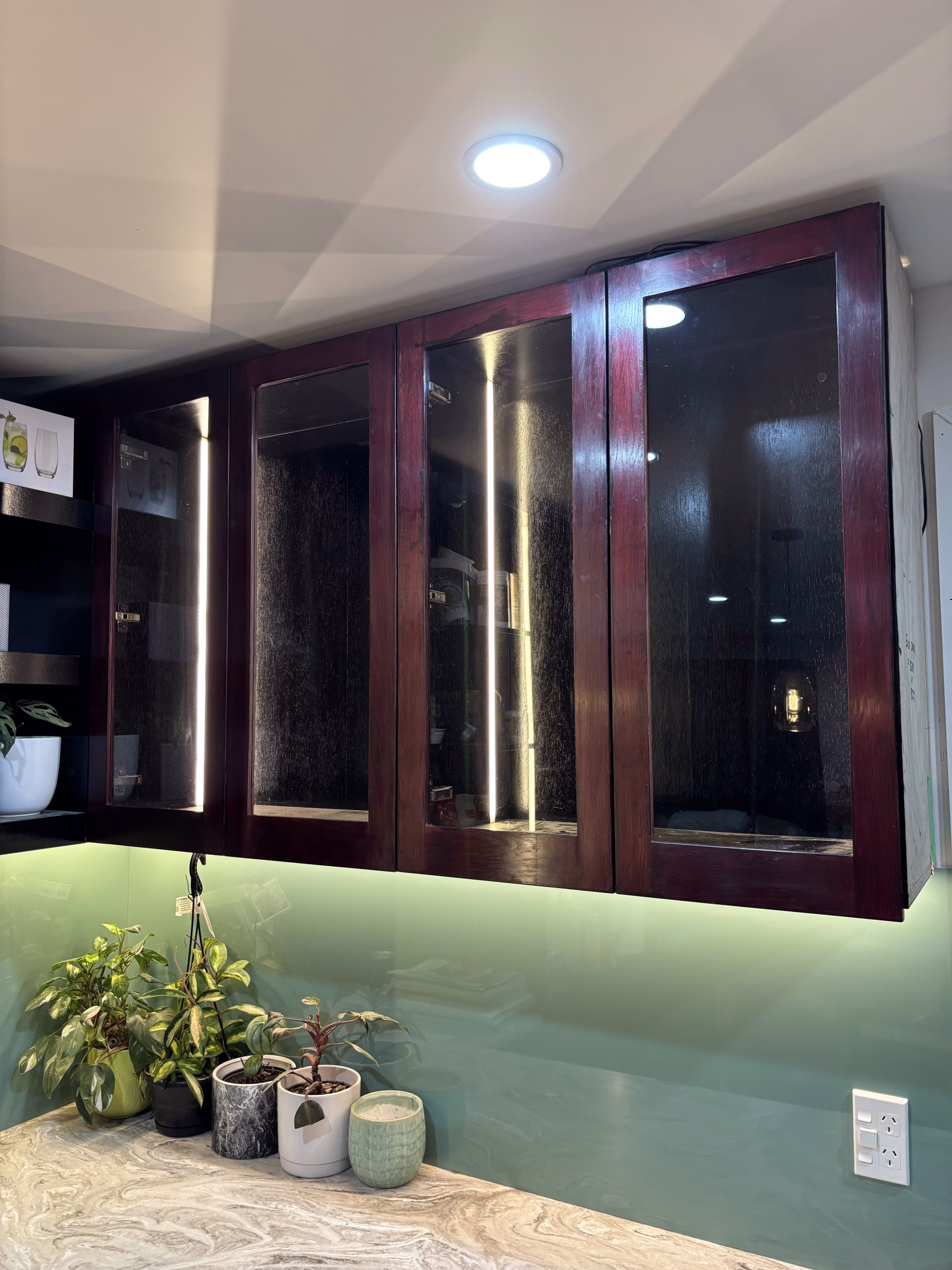 Dark timber wall cabinets with glass doors above a green splashback and marble benchtop with potted plants