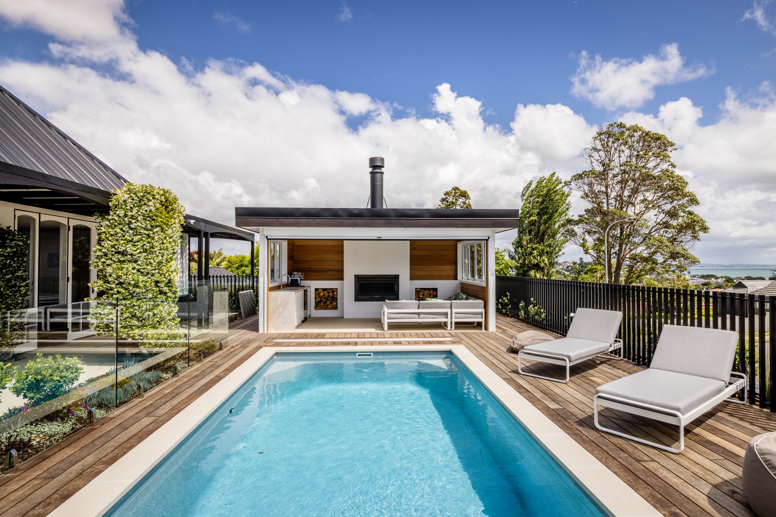 Outdoor pool and timber deck with a poolside cabana featuring a fireplace, outdoor seating, and sea views beyond