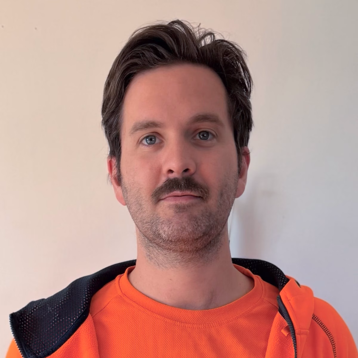 Man with dark hair and moustache wearing an orange high-visibility shirt and dark jacket