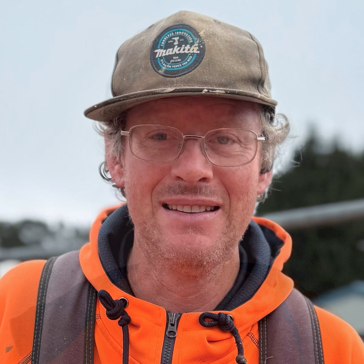Man wearing glasses, a Makita cap, and an orange high-visibility hoodie, smiling outdoors