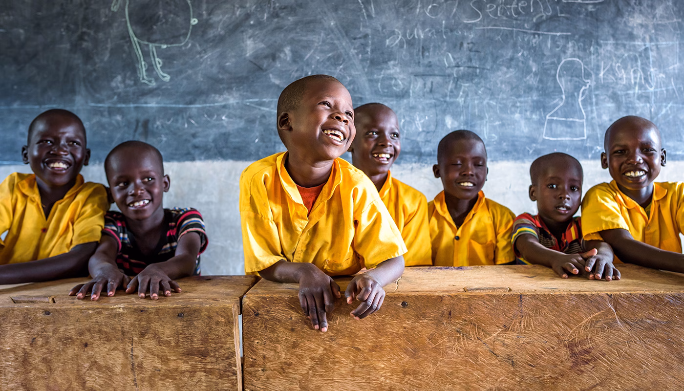 A group of smiling children in yellow school uniforms lean on wooden desks in a classroom with a chalkboard behind them