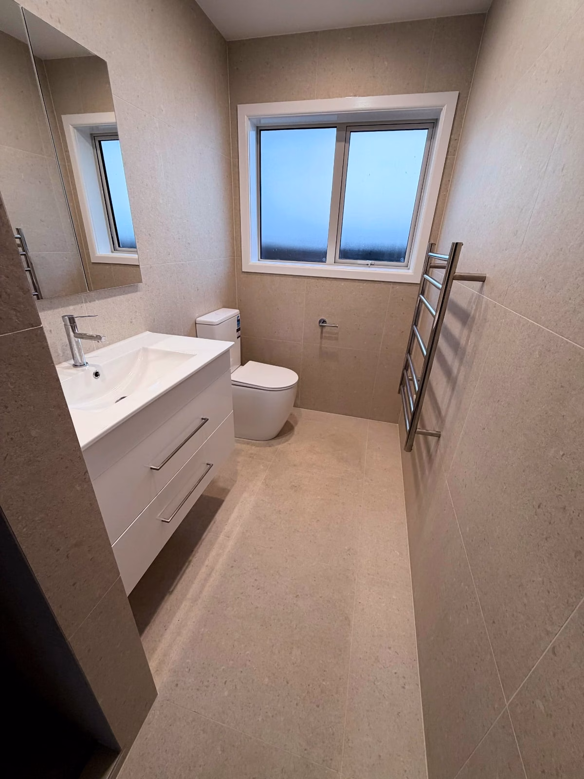 Narrow tiled bathroom with white vanity, mirror, toilet, window and heated towel rail