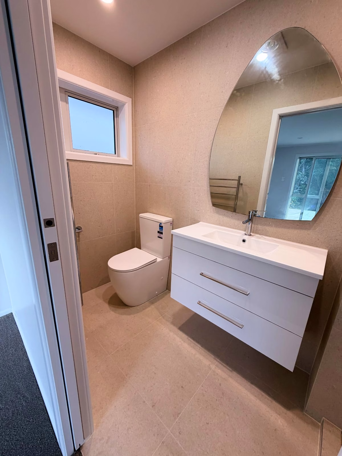Bathroom with beige tiling, a floating white vanity with two drawers, an arched mirror above the basin, a toilet, and a small window