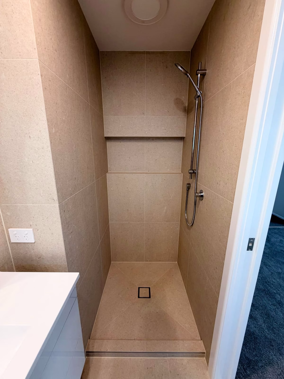 Tiled walk-in shower with beige stone-look tiles, built-in niche shelf, adjustable handheld showerhead, and square floor drain